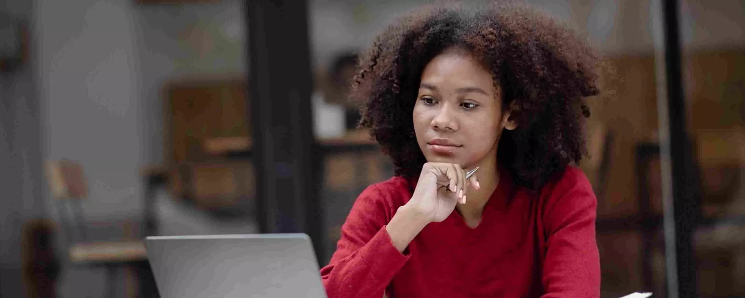 A young woman thoughtfully reviews her resume on a laptop, looking at the screen with her chin resting on her hand.