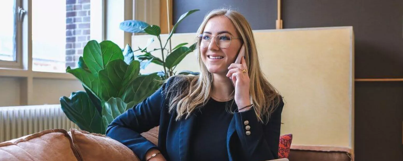 Eine Frau sitzt im Anzug auf der Couch vor dem Laptop und telefoniert