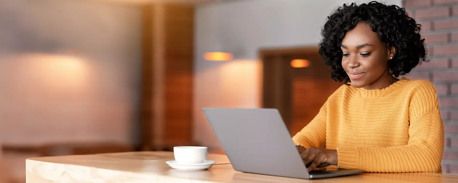 A smiling woman in a cozy yellow sweater works on her laptop at a wooden table with a cup of coffee nearby, in a warmly lit office.