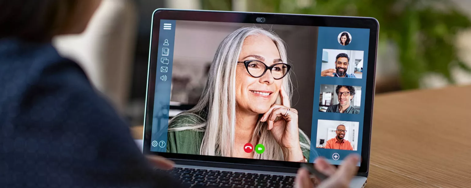 A smiling woman's face appears on a computer screen as an office worker interacts with participants in a virtual meeting.