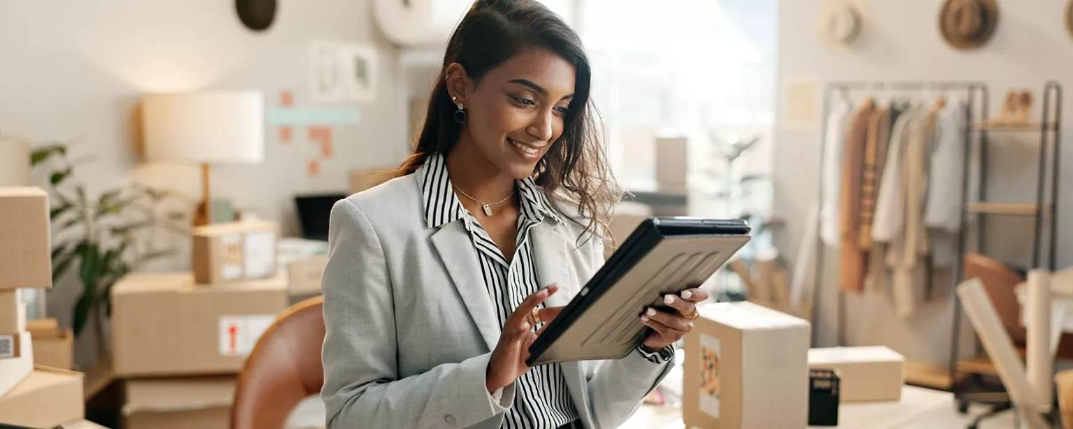 A young professional in a retail workspace smiles while using a tablet, surrounded by shipping boxes and clothing racks.