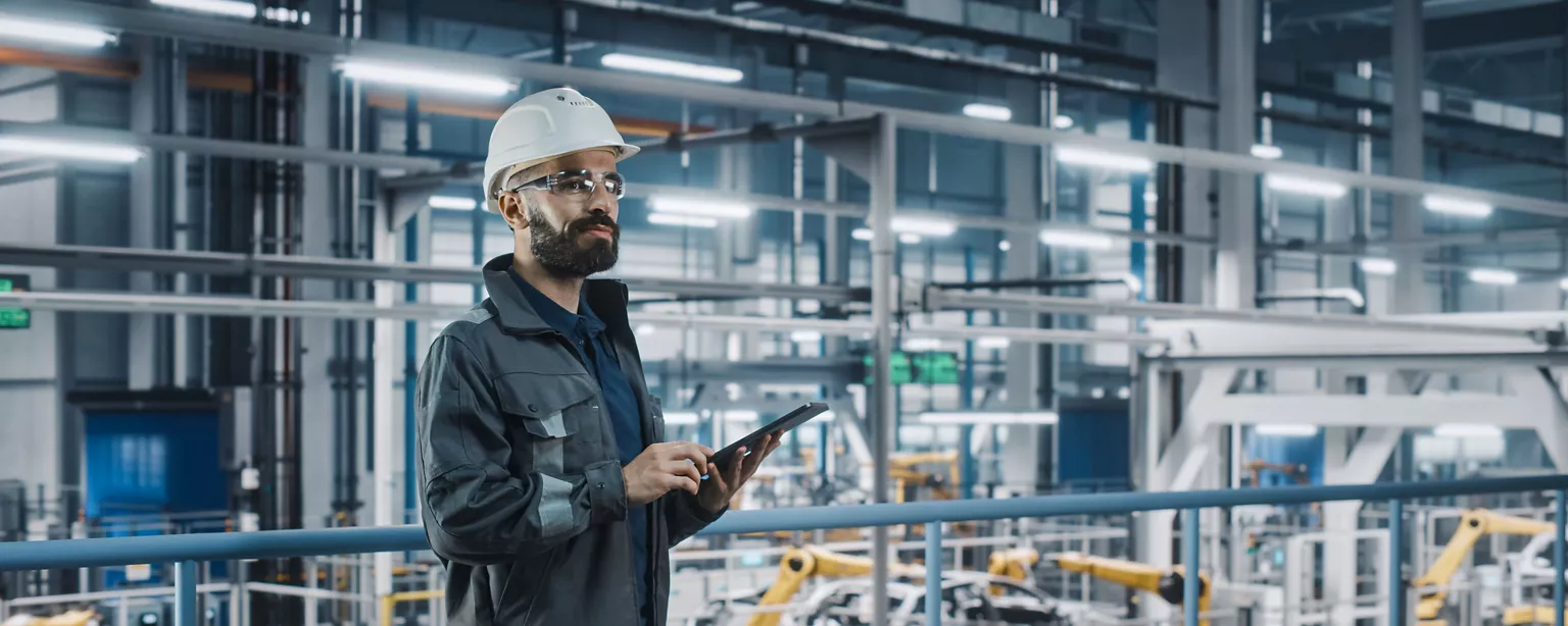 A male engineer wearing safety gear uses a digital tablet while overseeing operations in a modern, automated manufacturing facility.
