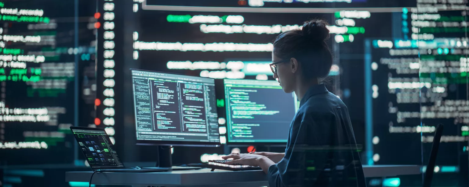 A woman sits at a desk in a dark, high-tech environment, focused on coding across multiple monitors displaying complex lines of programming code.