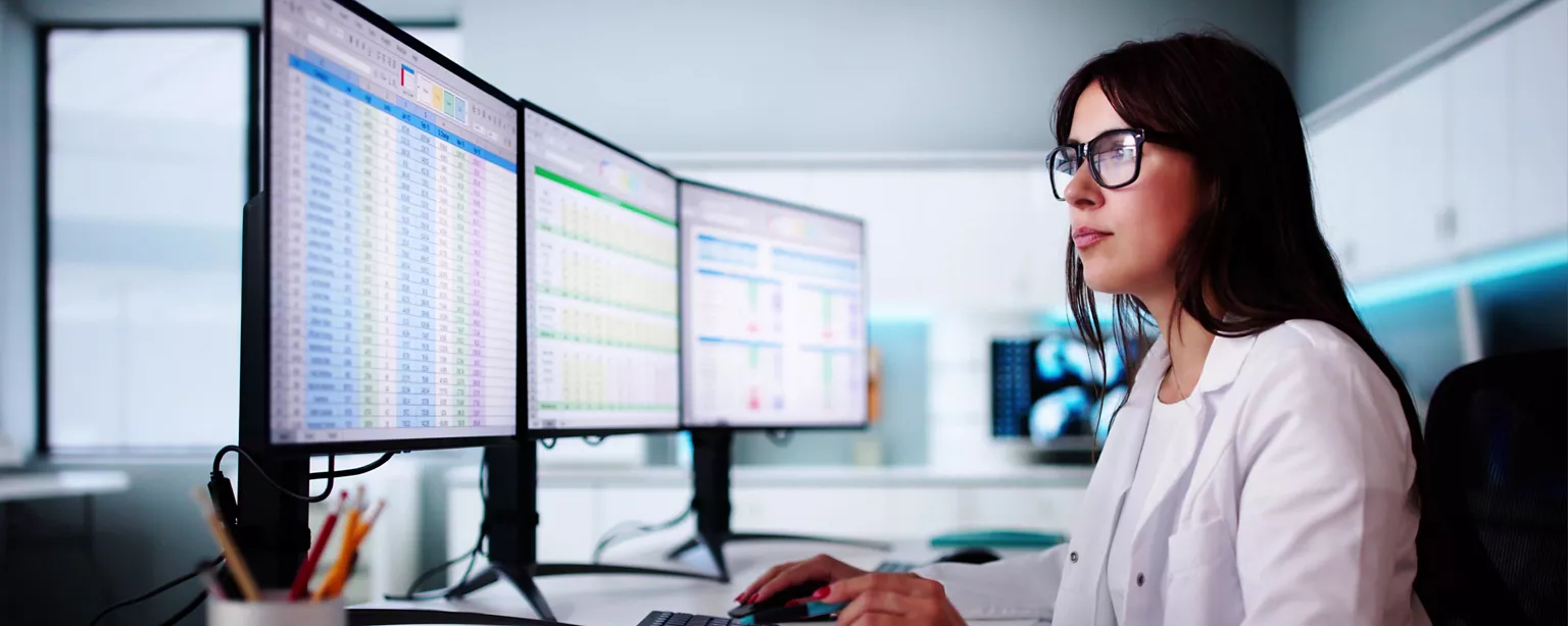 A healthcare professional in a lab coat analyzes data on multiple computer monitors in a modern medical office.