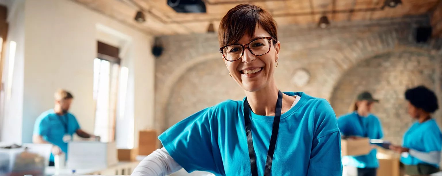 A smiling volunteer wearing a blue shirt and name badge stands in a nonprofit workspace with team members packing boxes in the background.