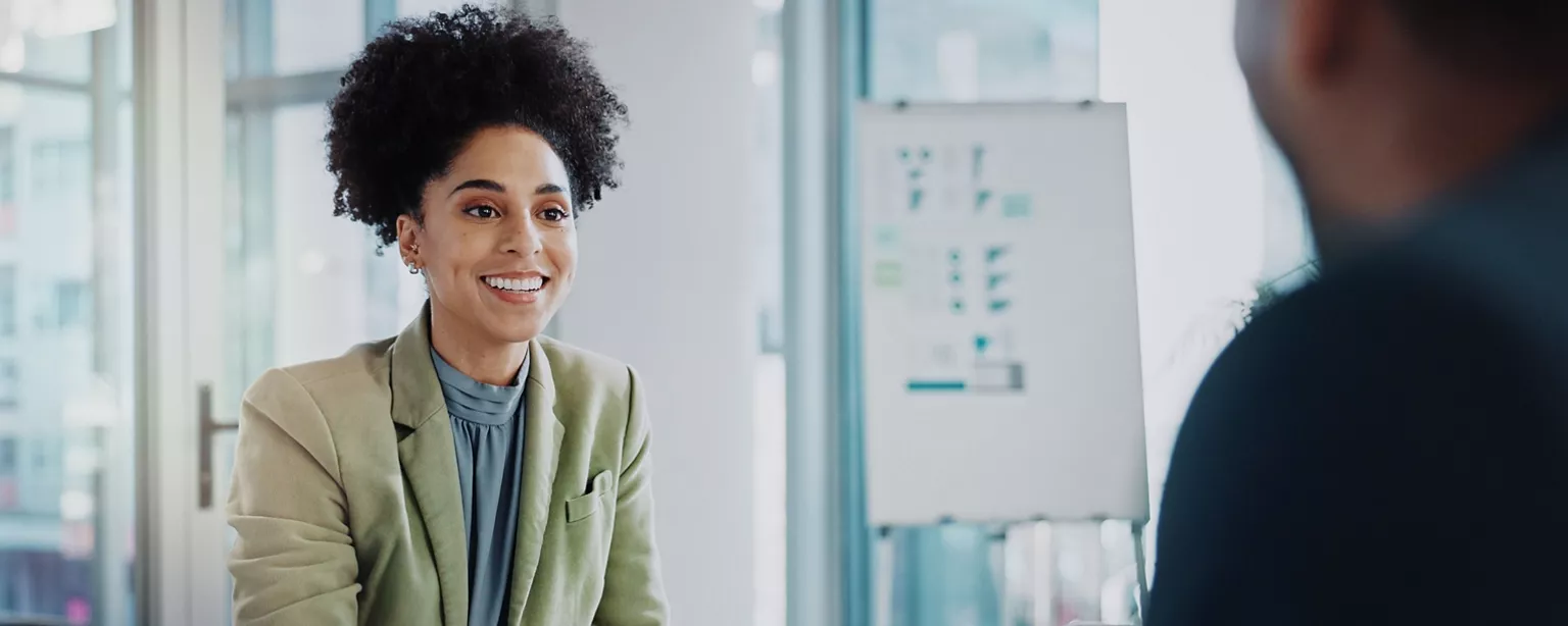 A smiling professional woman in a green blazer sits across from another person during an interview.