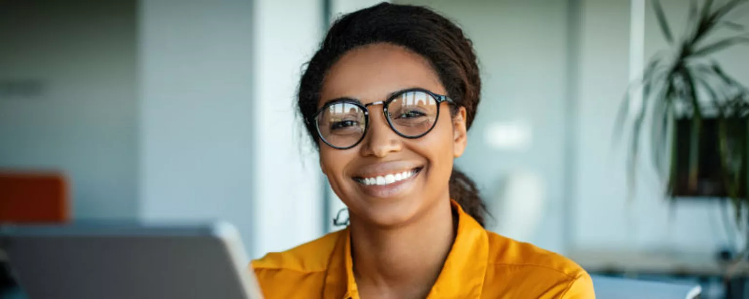 Smiling woman in glasses and yellow blouse sitting at desk in front of computer.