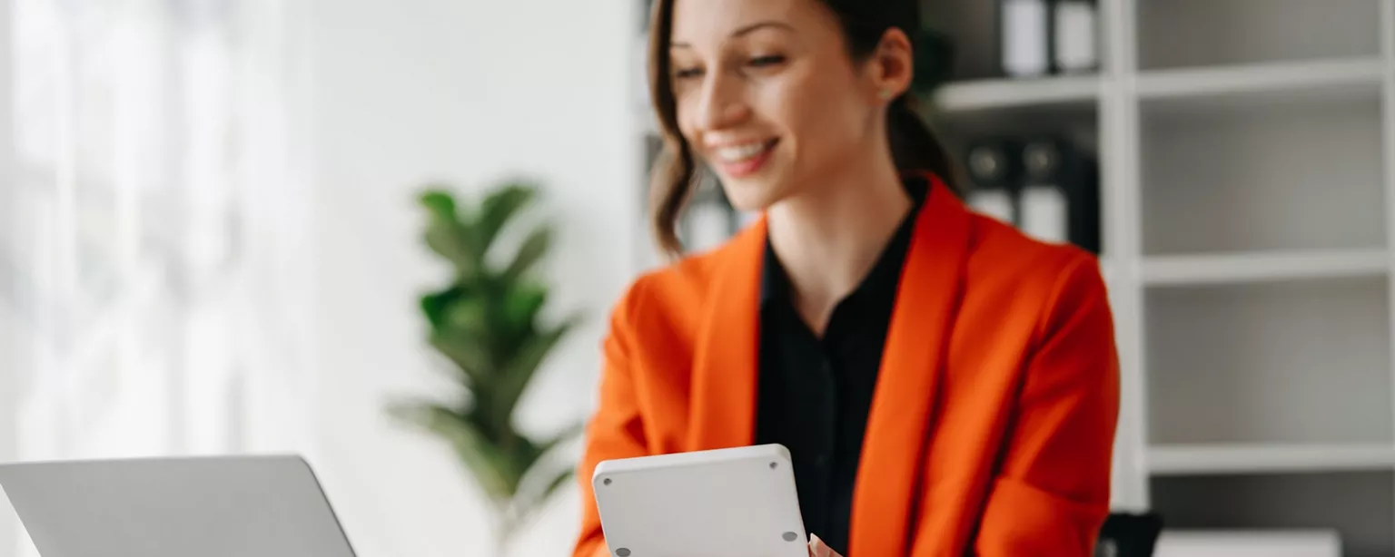 An accountant smiles at her desk as she uses a handheld calculator.