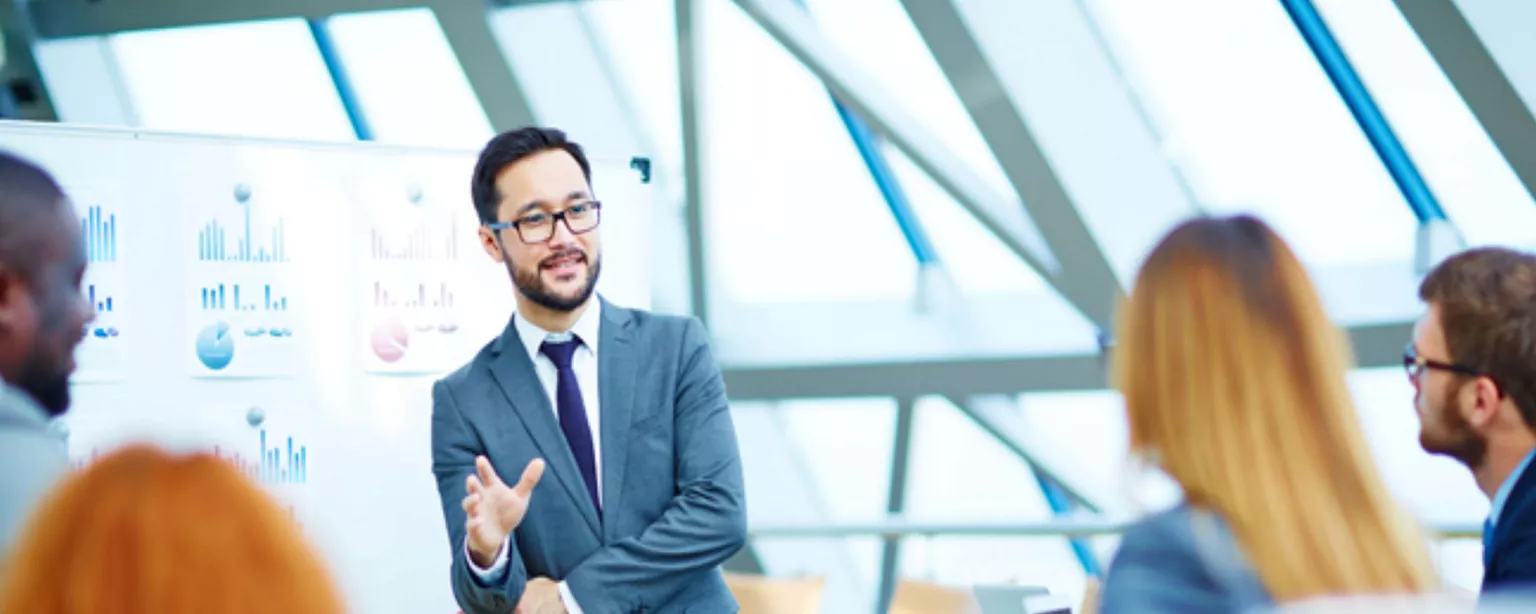 A financial executive standing in front of a white board with financial charts, addressing his team.