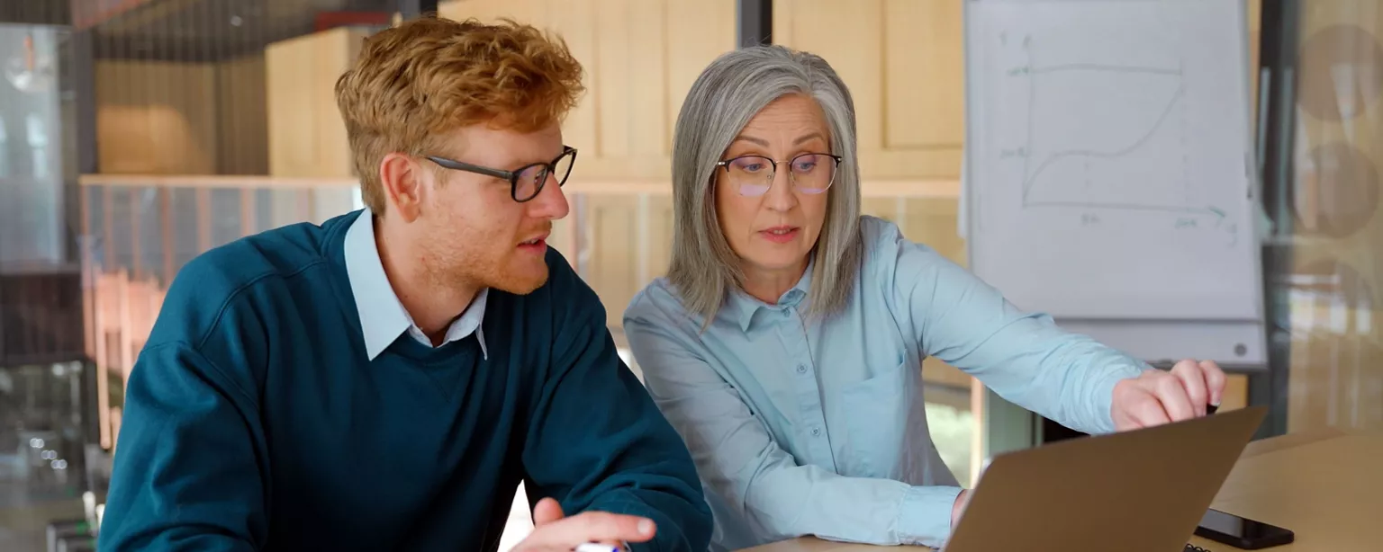 A newer employee watches and learns as his mentor, a more experienced female employee, instructs him on the next step.