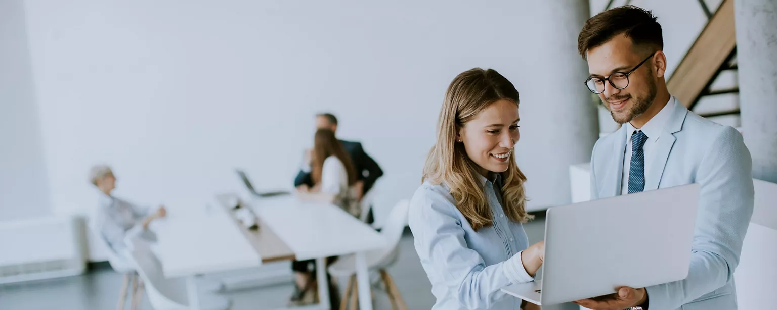 Two colleagues smile and discuss something on a laptop in a bright office while behind them other professionals chat at a conference table.