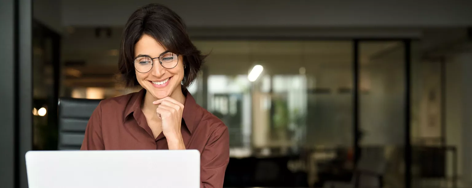 A smiling professional woman in glasses looks at her laptop while sitting in an office.