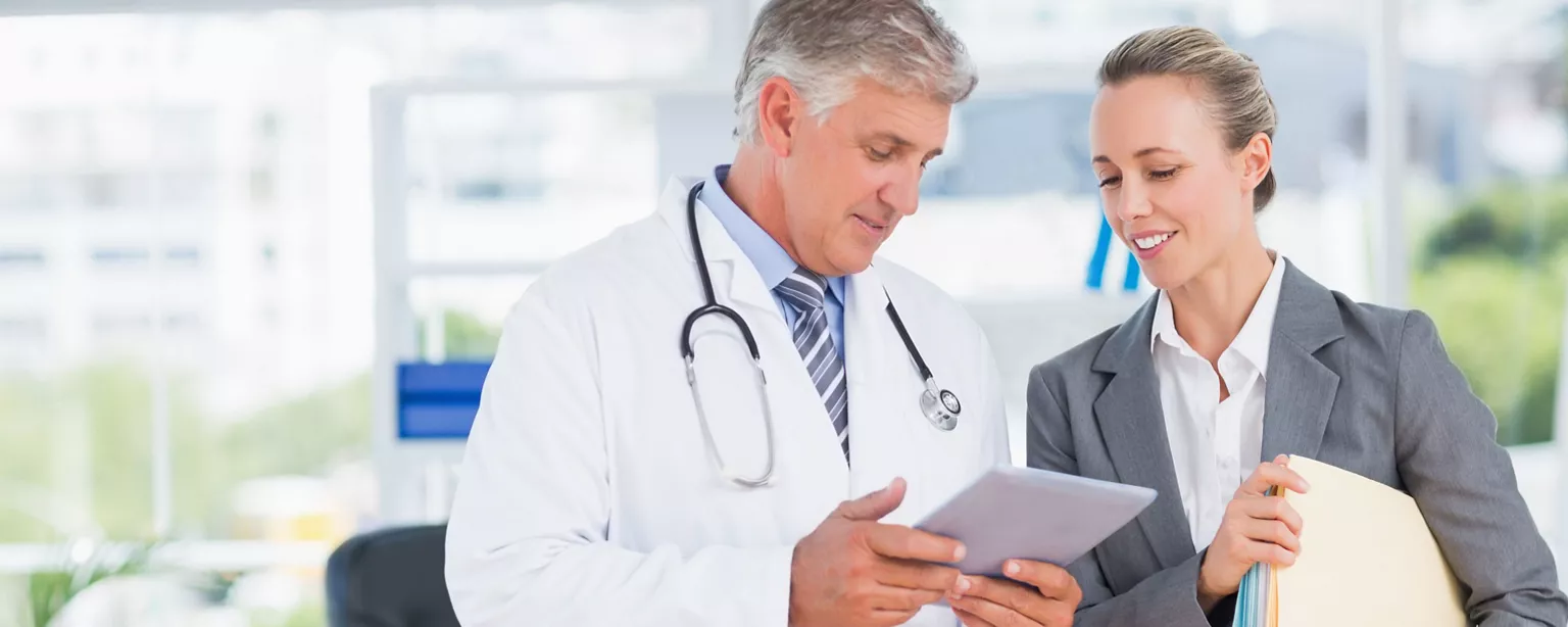 A healthcare administrator in a suit discusses information on a tablet with a doctor in an office with large windows.