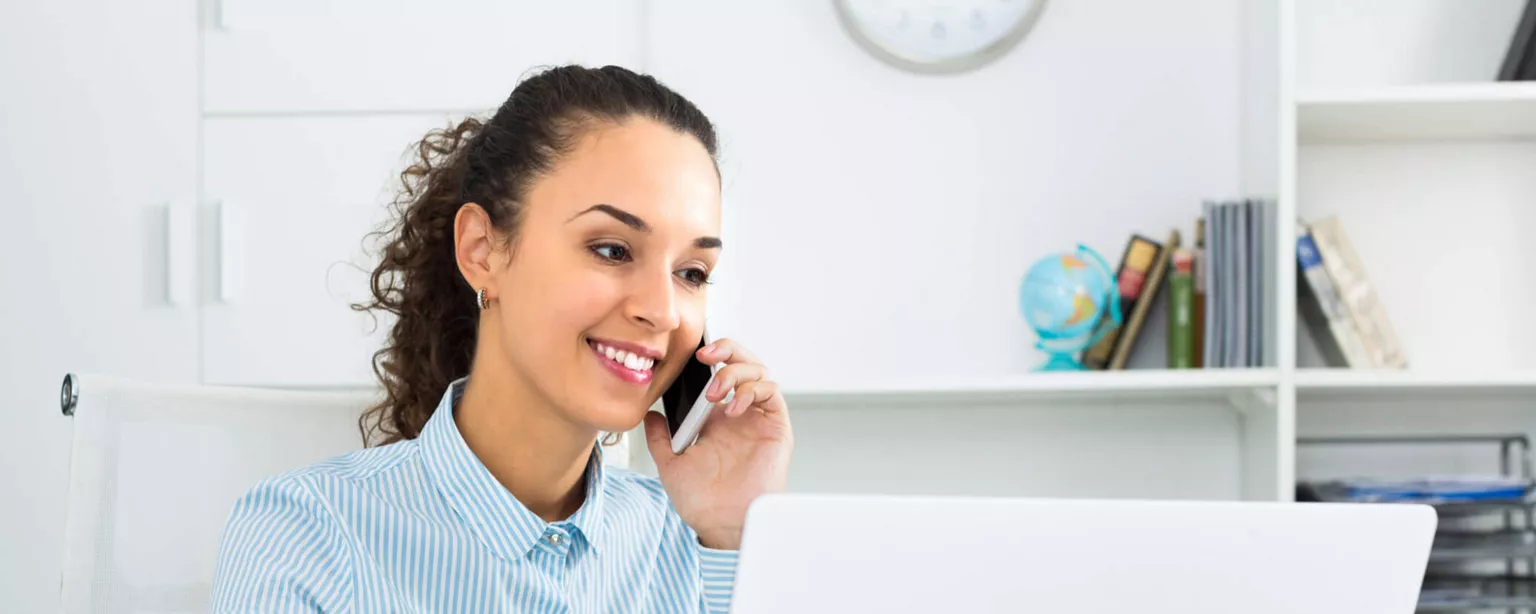 A woman with a phone to her ear smiles as she looks at her computer screen.