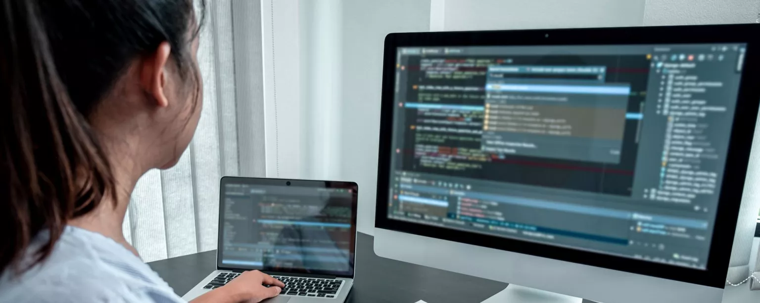 A young woman, who is a software engineer, is working on two computers and looking at a code database to try to solve a development problem.