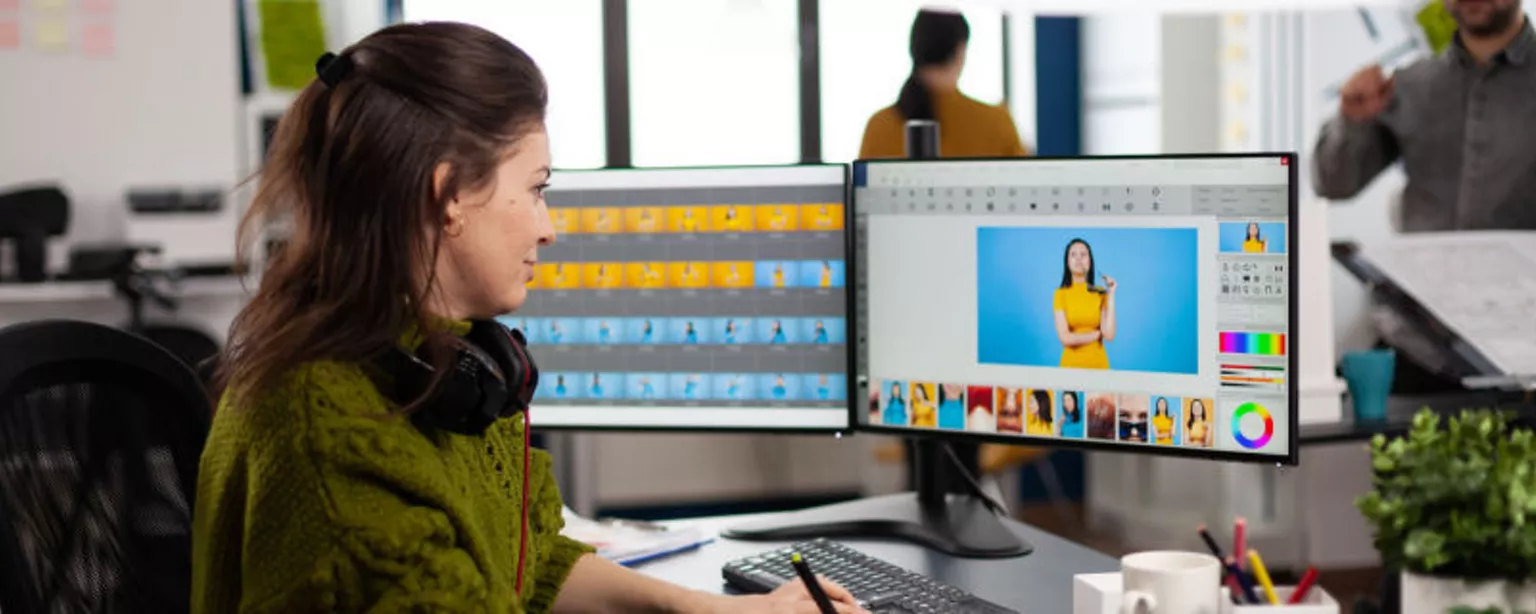 A production artist works with colorful images on two computer screens in the office.
