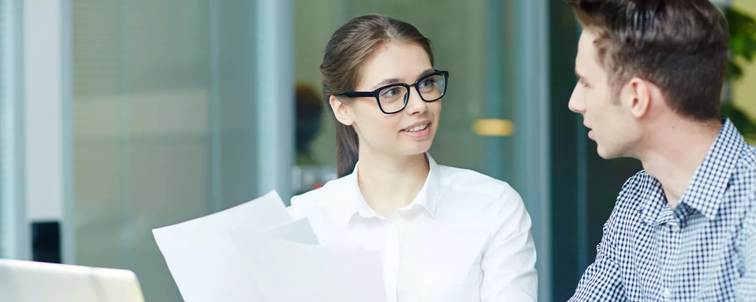 A woman in glasses is asking for feedback from a colleague while holding a stack of papers in a professional office.