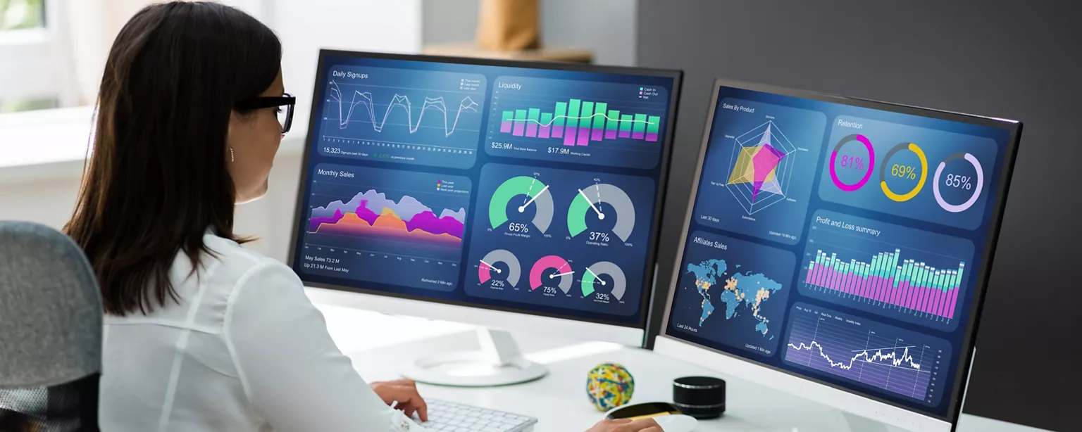 A business analyst evaluates the data on two large monitors as she works at her desk.