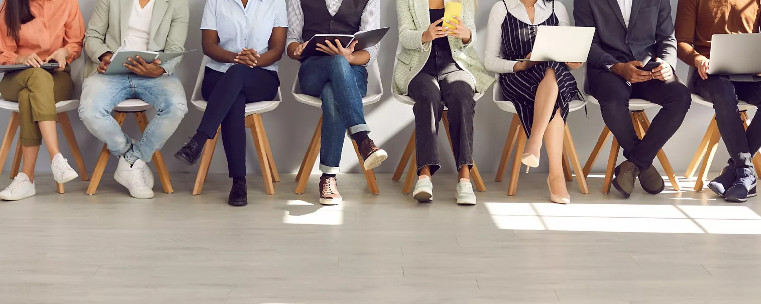 A row of job candidates seated with laptops and resumes, waiting for interviews in an office.