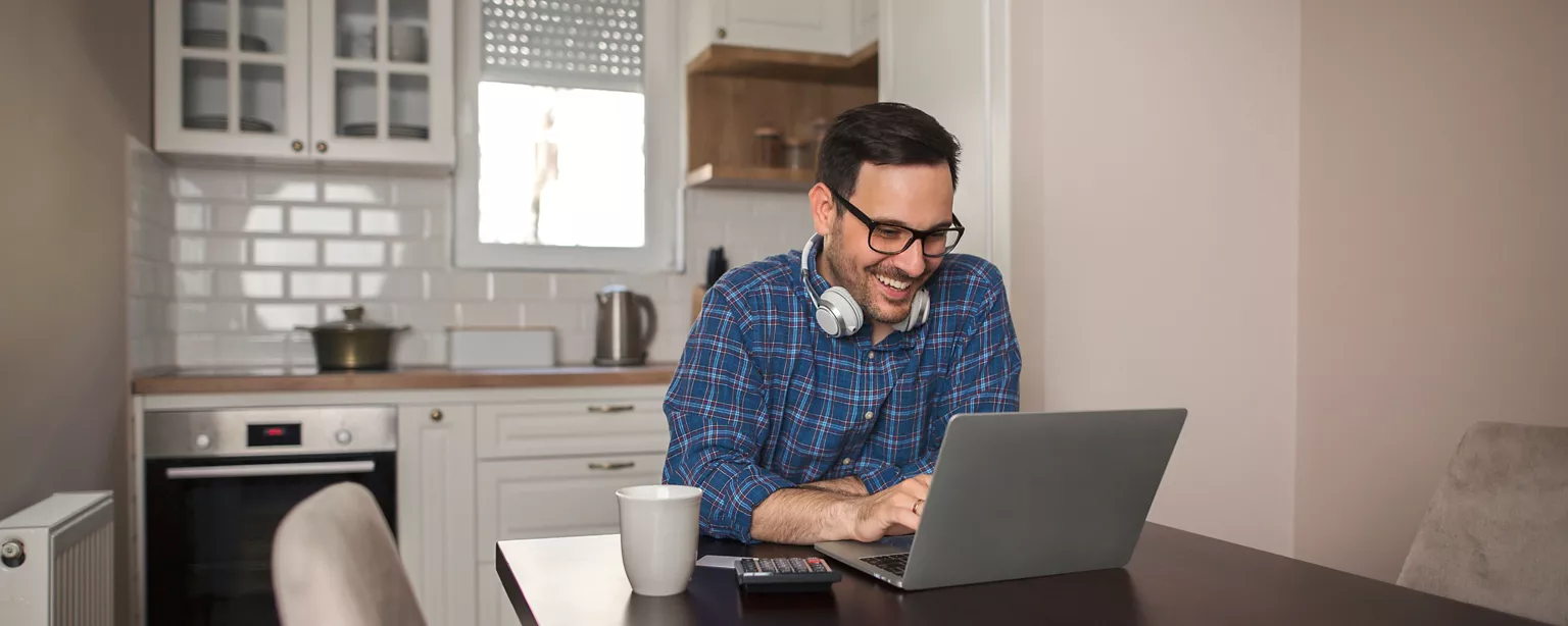 A cheerful man wearing glasses and headphones works on a laptop in his kitchen, probably asking about the hiring timeline.