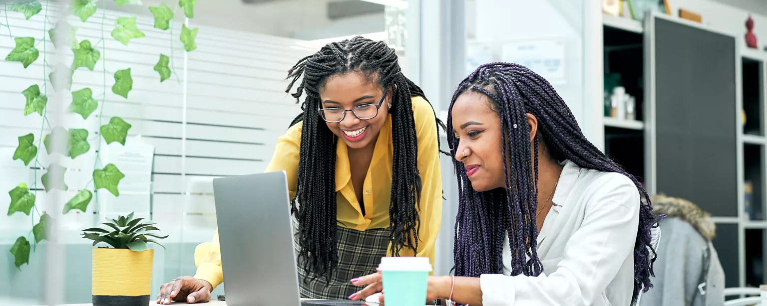 A woman seated at a desk gets assistance from a coworker helping to upskill her existing skills for a skills-adjacent role. 