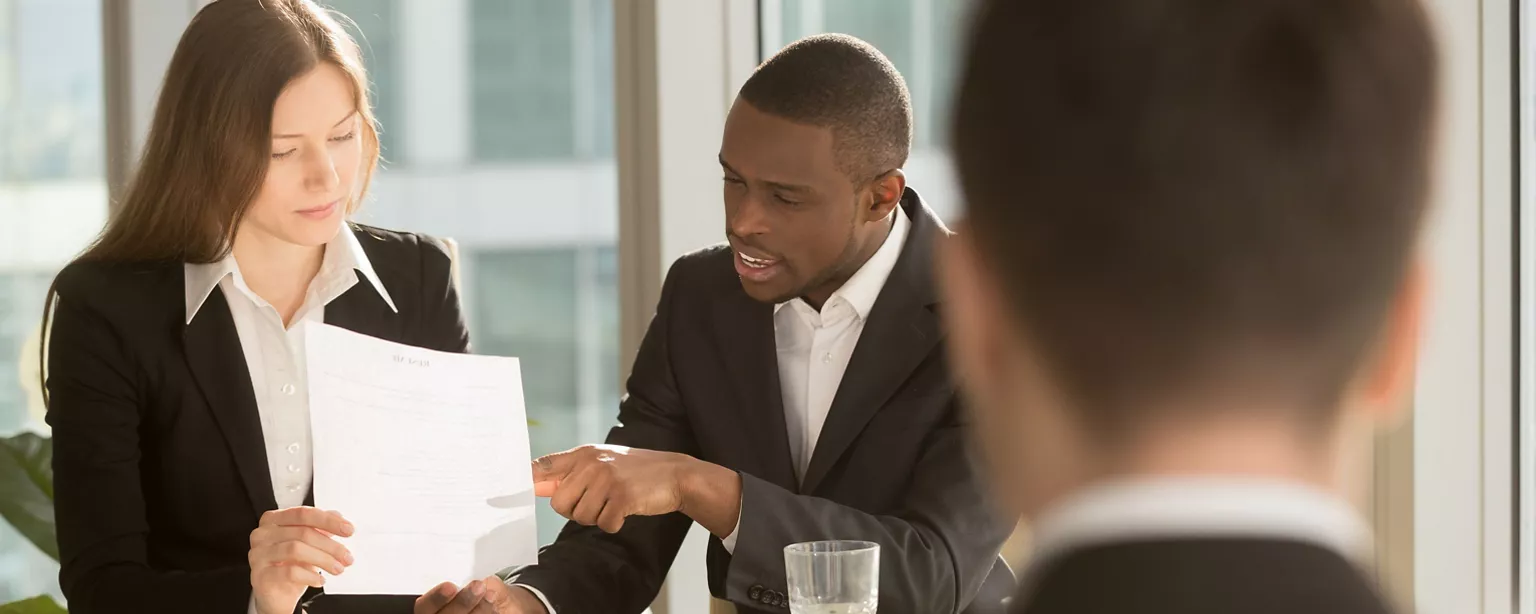 Hiring managers consult at a table as one holds the resume of a job candidate, who is pictured from behind in the foreground.