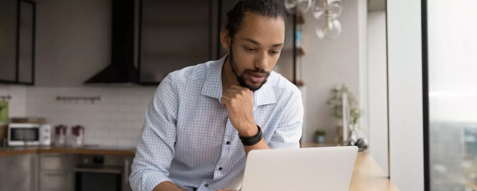 A man strikes a thoughtful pose as he looks at his laptop while writing an emailing requesting a raise.