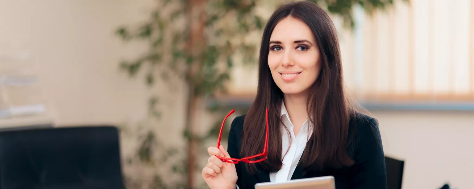 An executive assistant holds a pair of red glasses as she  works in an office.