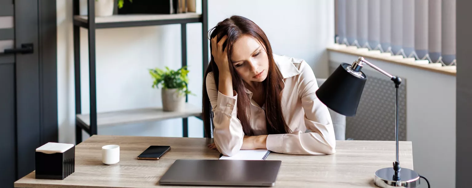 A young woman sits at an office desk, looking at her closed laptop with an expression of disinterest.