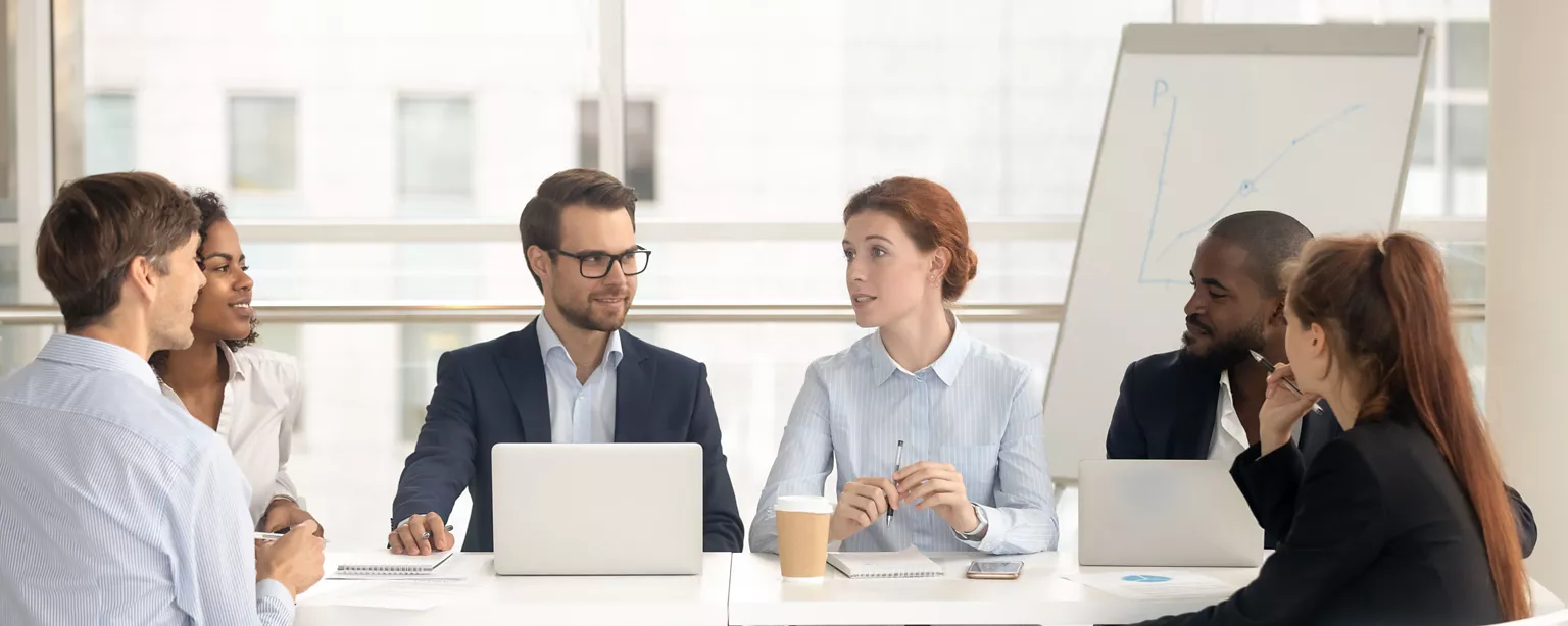 A group of professionals engaged in a team meeting around a conference table, with laptops, notebooks, and a presentation chart in the background.