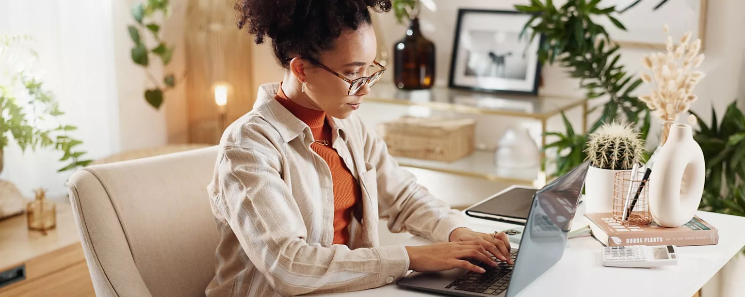 A copywriter works on a laptop in a bright, plant-filled home office with a stylish and organized desk.