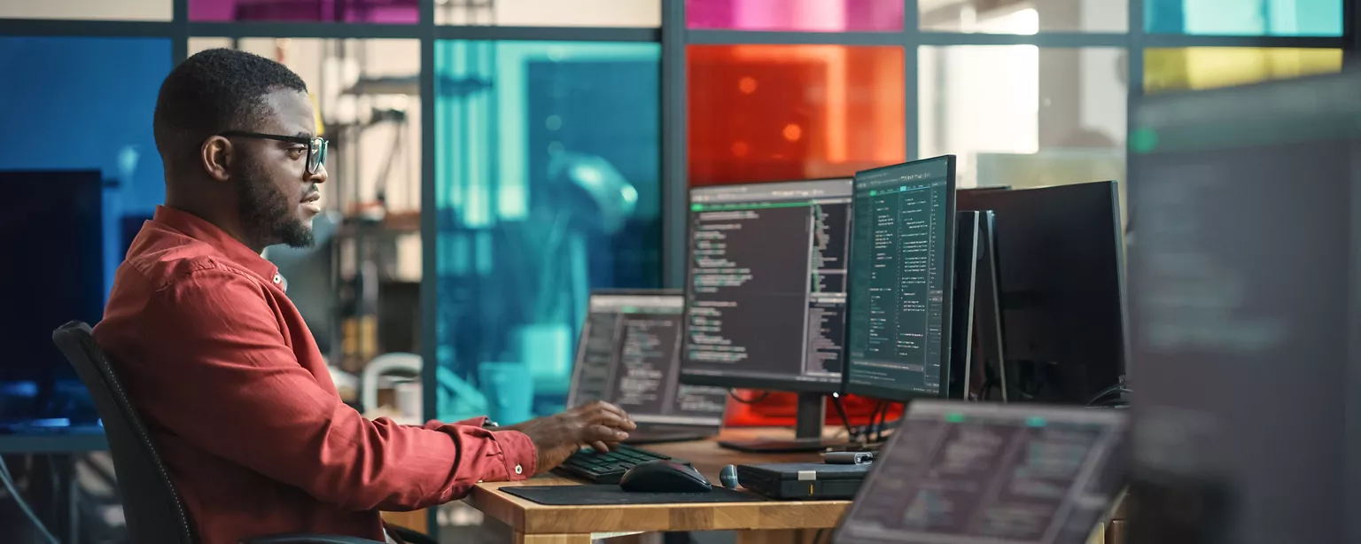 A data scientist wearing glasses and a red shirt works at multiple computer monitors displaying information in an office with colorful glass panels.