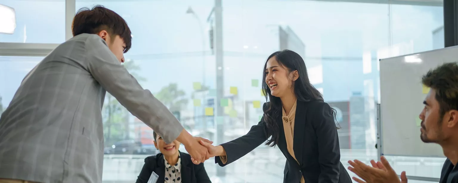 A professional woman smiles and shakes hands with a colleague during a friendly introduction in a workplace meeting.