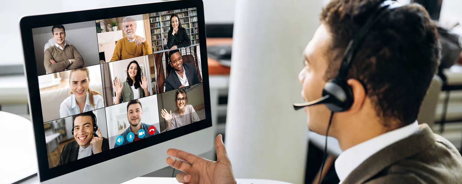 A man wearing a headset in an office environment confers with colleagues on a computer video screen, many of whom are working from home as part of a hybrid work model arrangement. 