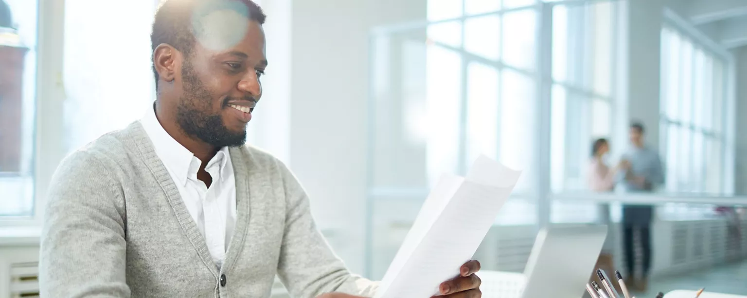 A professional reviewing a resume at his desk in a bright office and two coworkers talking in the background..