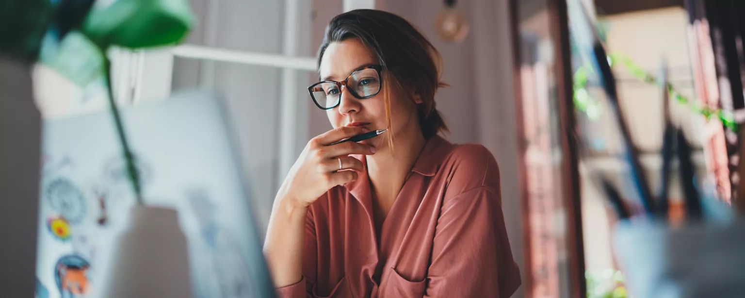 Woman in glasses sitting at desk in dark red top with hand at chin and looking at computer