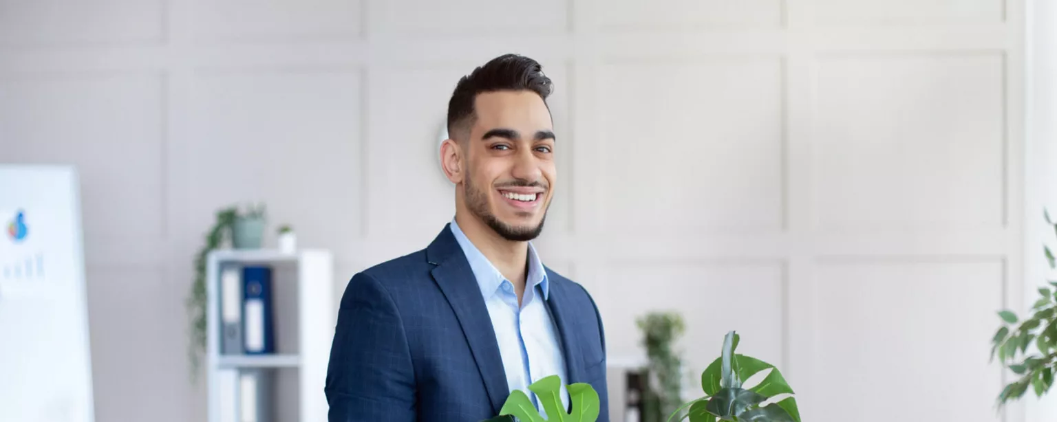 Smiling businessman in an oxford shirt and blazer moving out of his office, and carrying a box of office supplies and a plant.