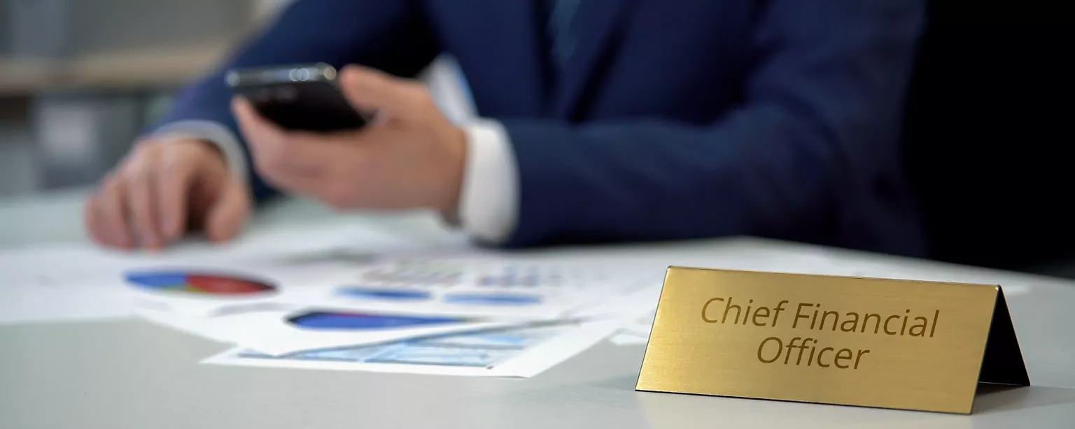 A CFO's hands are visible as he sits at his desk reviewing numbers on a calculator, with the words "Chief Financial Officer" seen on a brass nameplate.