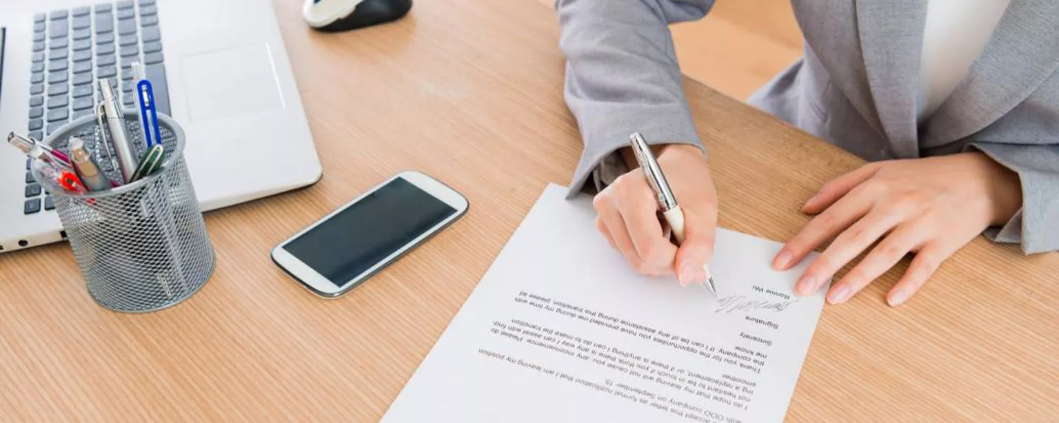 How to Write a Resignation Letter — Person signing resignation letter at desk with computer and phone