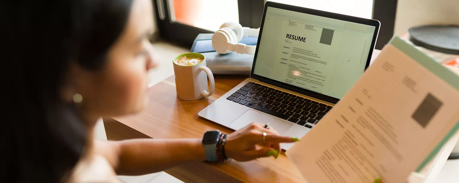 A woman reviews a printed resume while referencing a digital resume on her laptop at a desk with a cup of coffee.