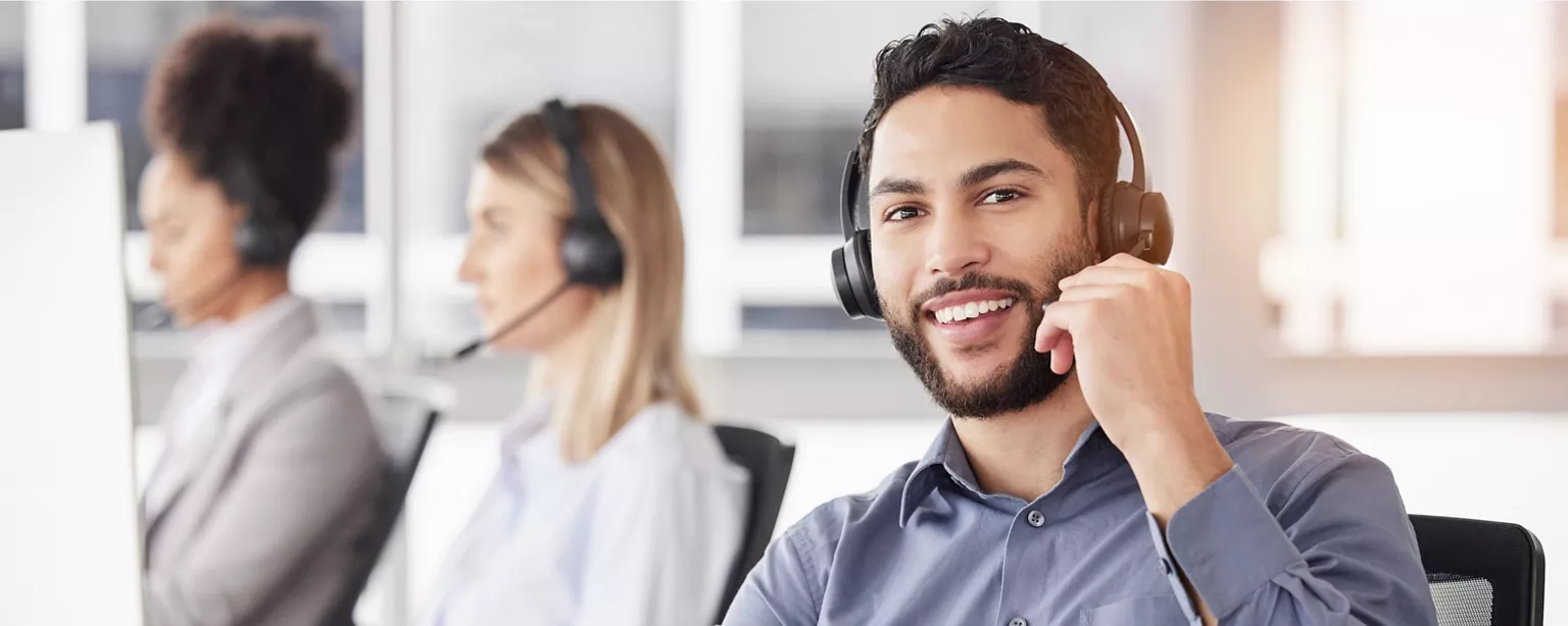 A professional young man wearing a wireless phone headset smiles while sitting with his fellow IT Help Desk coworkers.