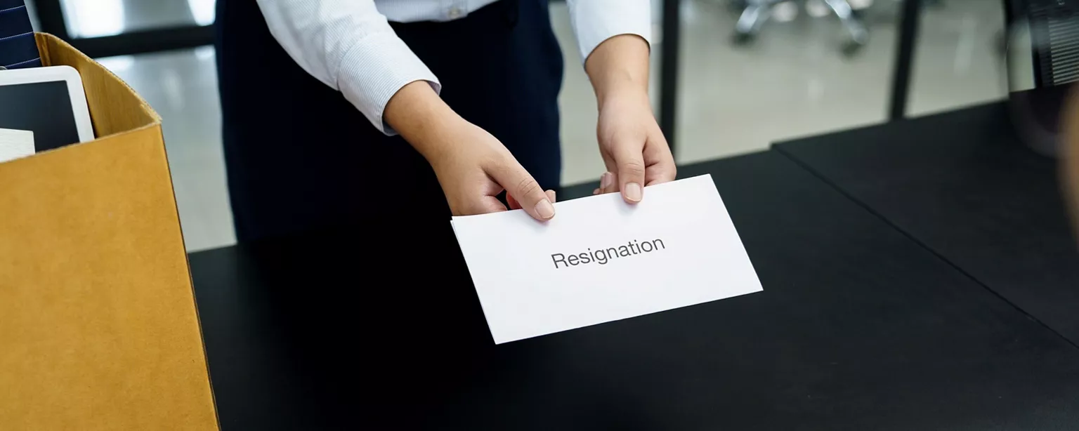 A person uses both hands to present an envelope labeled “Resignation.” They are standing in front of a black desk; a cardboard box with office supplies is next to them.