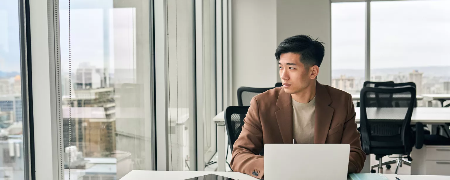 Man in office sat at his desk, looking away from his laptop, to the window