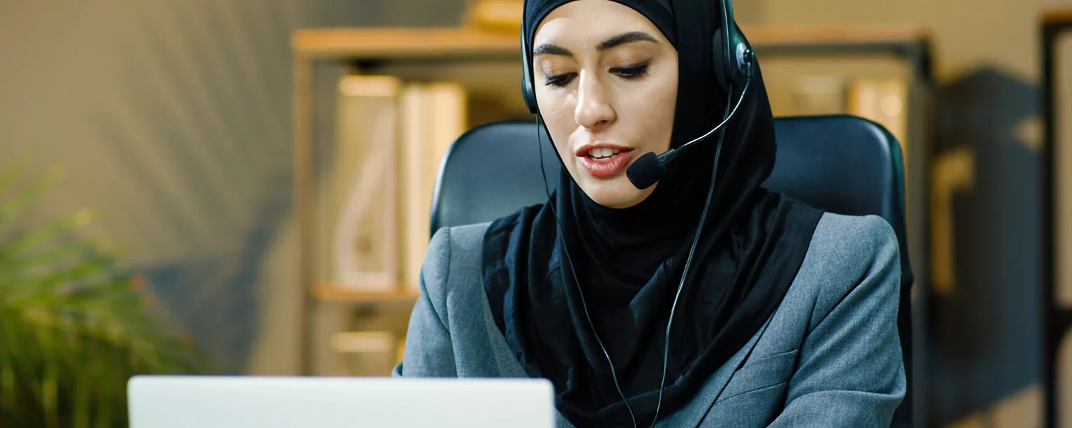 Middle Eastern woman wearing hijab at a computer, with a headset around her ears, speaking into the headset mic while typing.