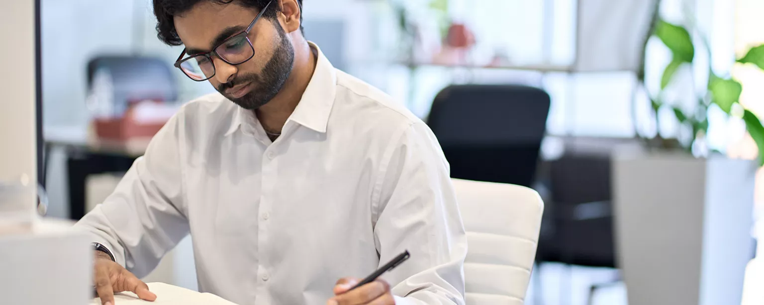 South Asian man with black hair, beard, and glasses with a pen in his left hand reading over white paper sheets at his desk.