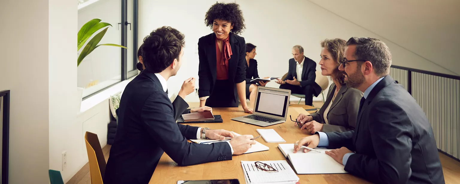 Group of professionals wearing business suits around a desk, papers between them, paying close attention to a frizzy-haired Black woman who is the only person standing.