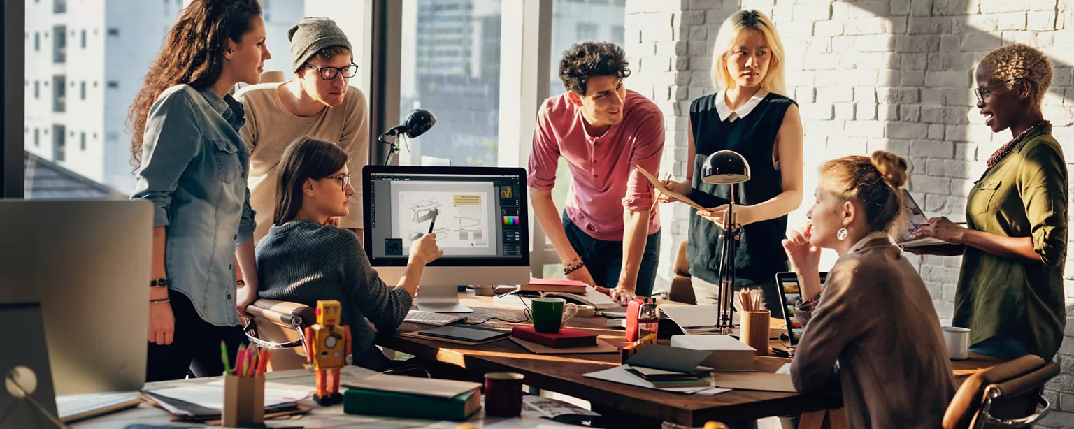 Group of creative professionals of diverse backgrounds and in diverse clothing, in a naturally lit room with windows along one side, paying attention to a Black woman with closely-cropped, dyed blonde hair speaking on the right.