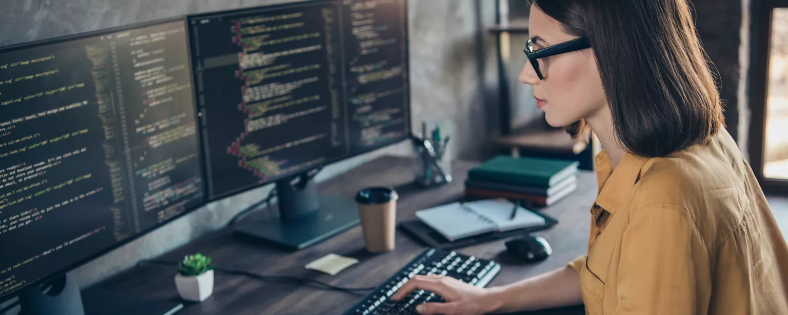 White woman in yellow shirt with long brown hair and glasses looking at lines of code on two computer monitors.