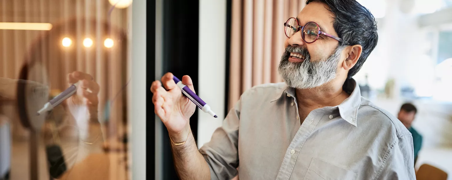 A mature entrepreneur with a greying beard and glasses in a cafe, holding a pen in his right hand and smiling at his reflection in a sliding door, as if he's had a winning idea.