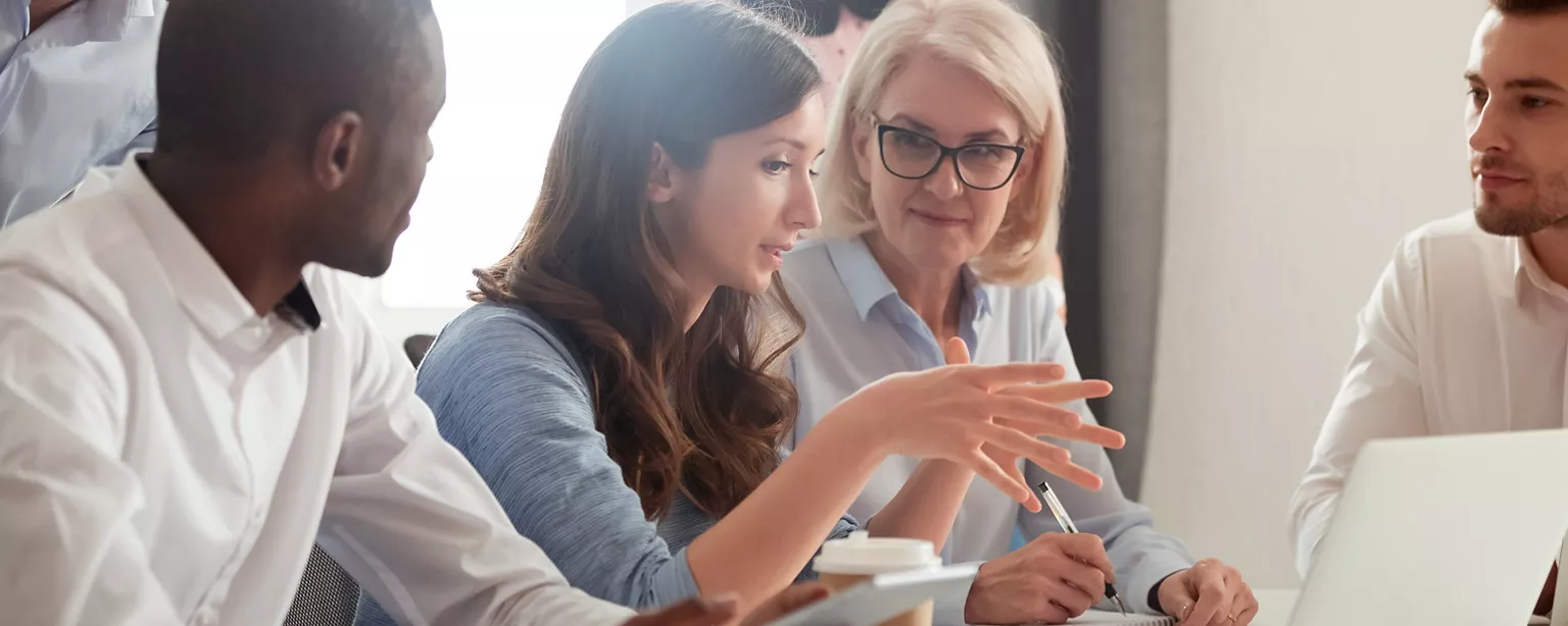 A group of co-workers are engaged in a collaborative discussion around a table, with a laptop and notepad visible.